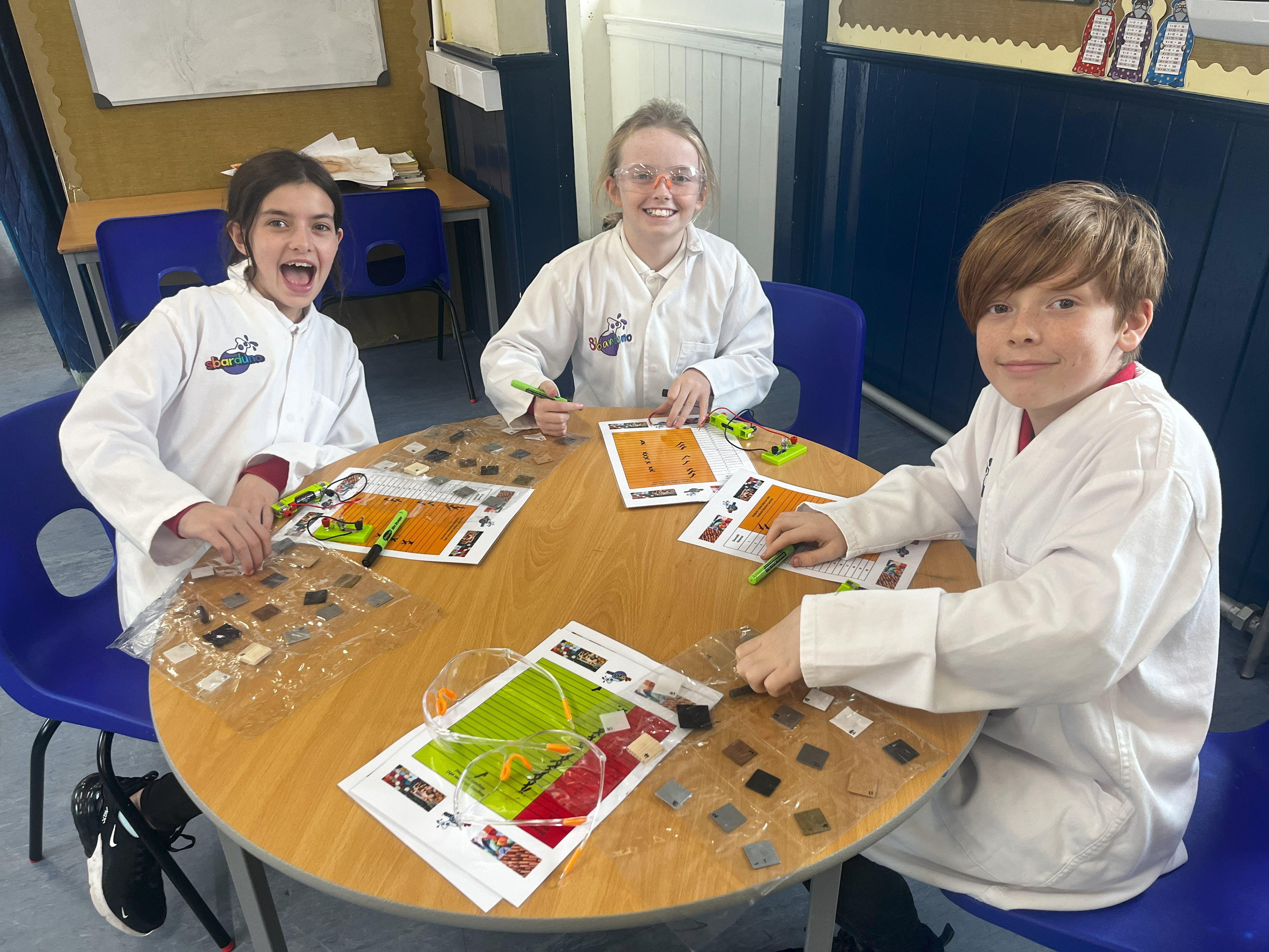 Three primary school children in Wales sat around a table wearing white lab coats, carrying out a science exercise.