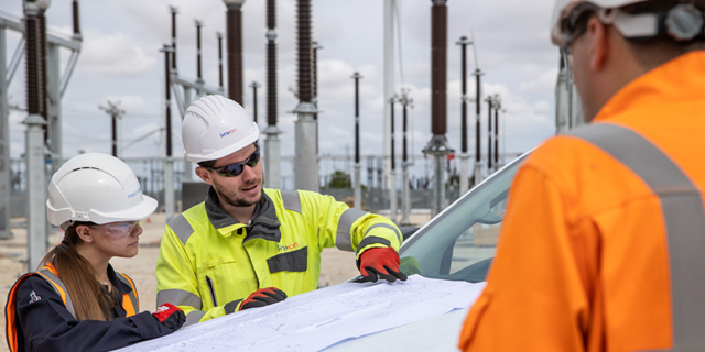 Three engineers wearing PPE looking at a drawing on the bonnet of a car