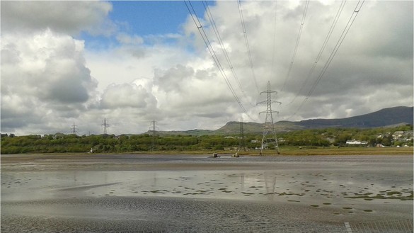 A stretch of water, sand, and land from Snowdonia with grey clouds and electricity pylons in view 