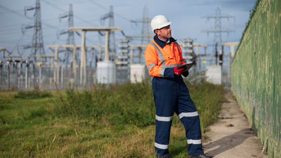 Engineer wearing PPE and holding a tablet inspecting a fence by an electricity substation