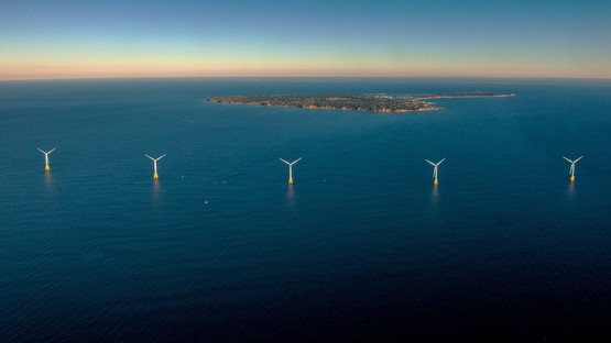A seascape of dark blue ocean with an island in the background and several wind turbines appearing up from the water