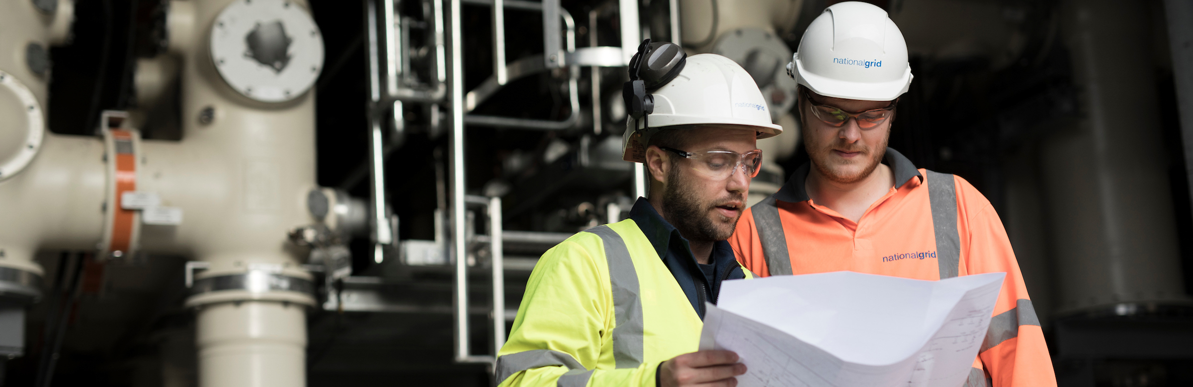 Two National Grid workers looking at a sheet of paper with an industrial framework in the background - About us