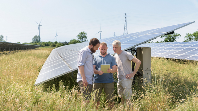 Three men standing in front of solar panels in solar farm field, with wind turbines and pylons in the background