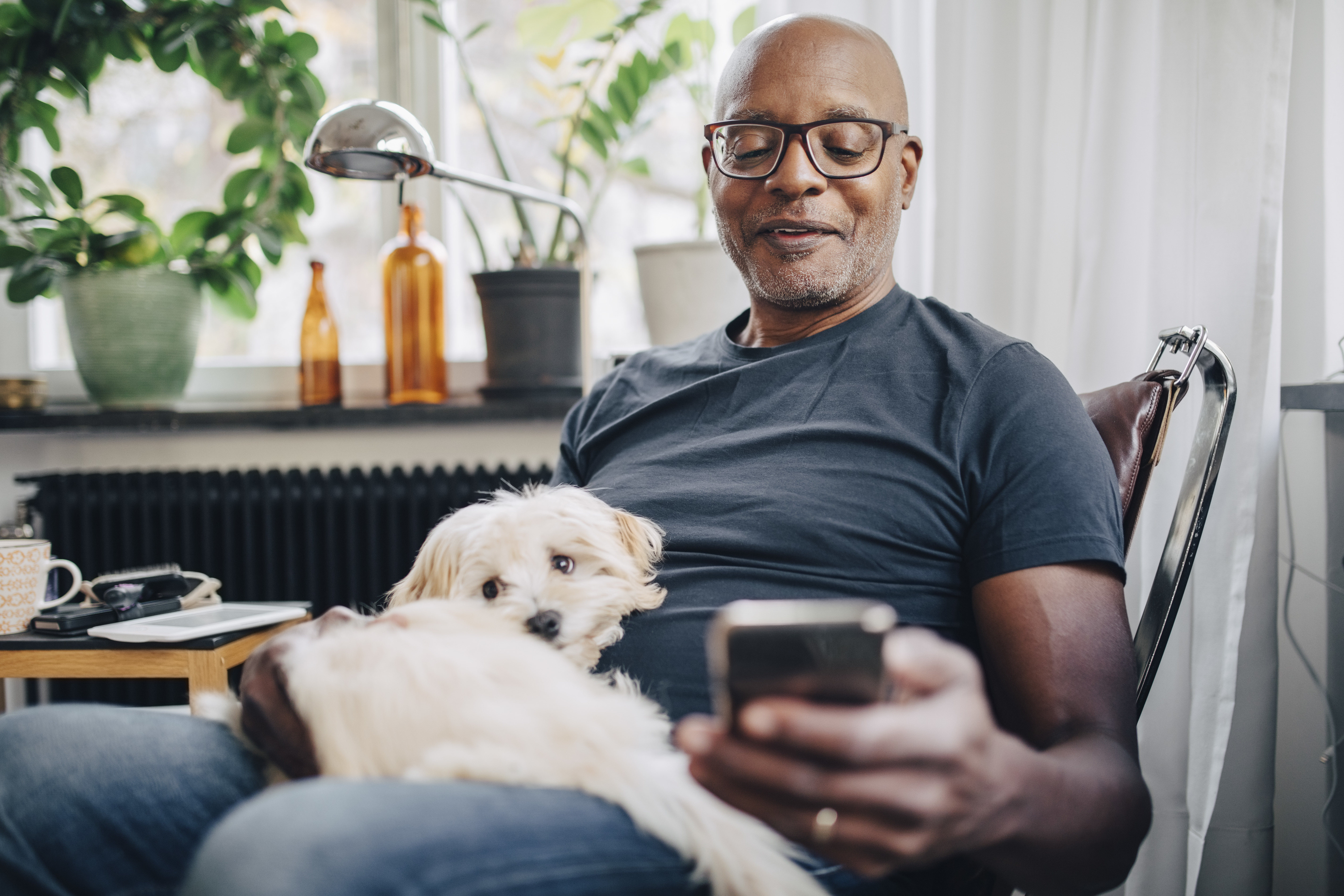 Smiling retired senior male using smart phone while sitting with dog in room at home