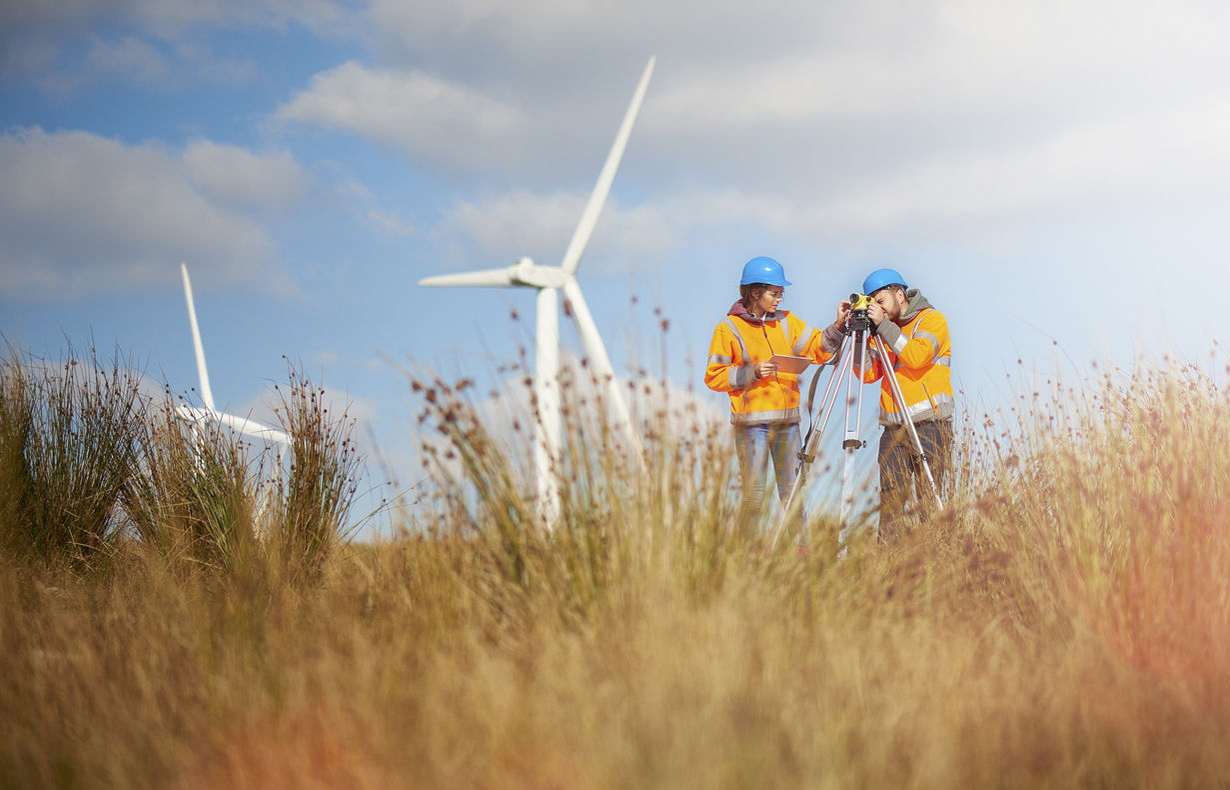 Man and woman standing in grassy field in front of windfarms, wearing hard hats and high-vis jackets with a survey camera - used for the National Grid story 'Now is the time to recruit a Net Zero Energy Workforce to power a greener future'