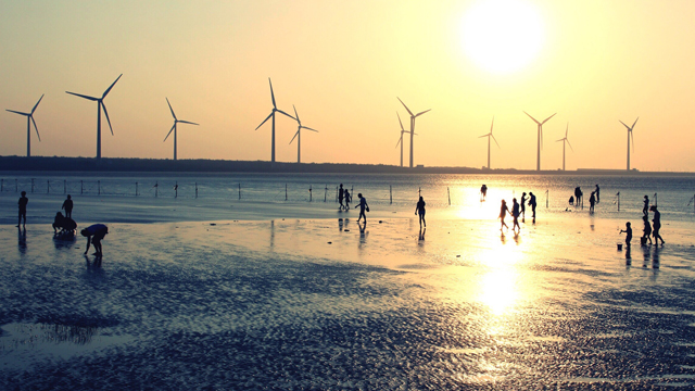 Silhouettes of people on a beach at sunset with wind turbines out at sea in the background