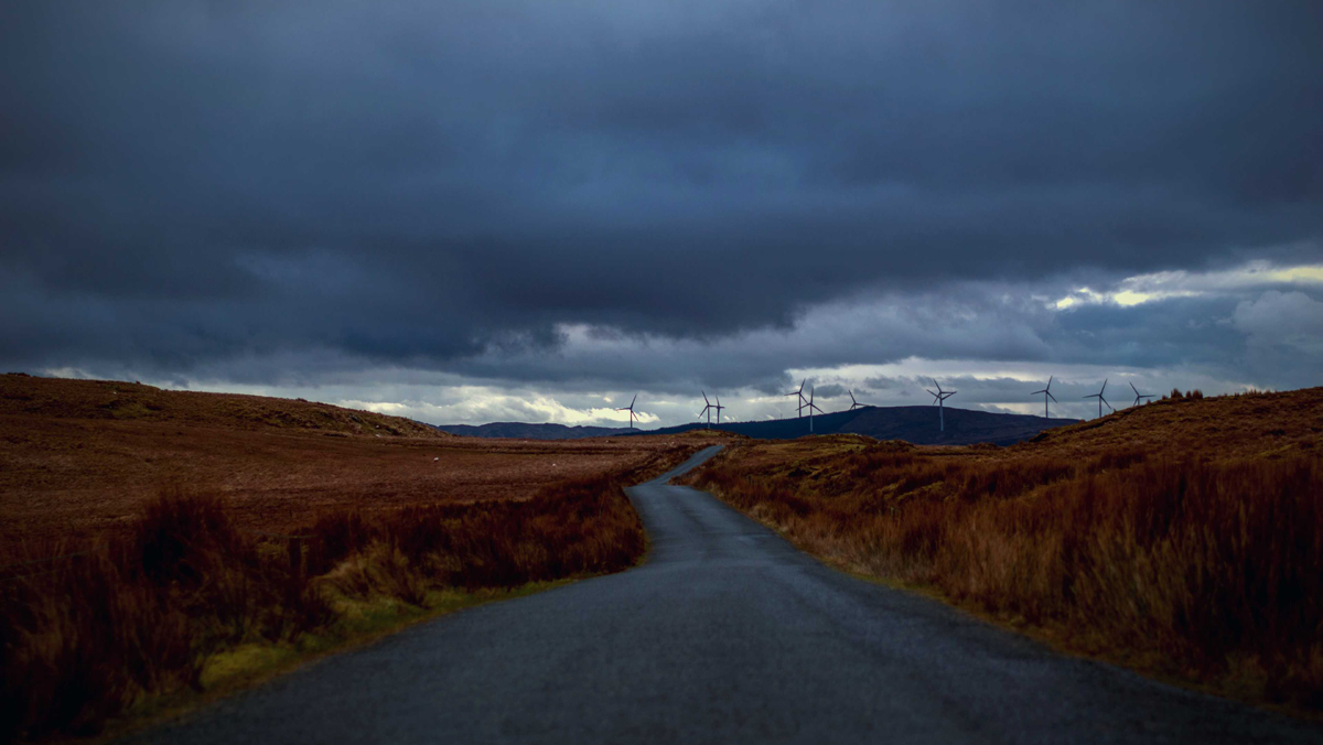 Empty road between brown moors under a stormy sky with wind turbines on the horizon - used for the National Grid story 'Riding the storm: how the ESO control room handles severe weather'