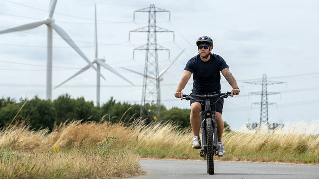 Man wearing helmet, shorts and t-shirt cycling on country road with wind turbines and electricity pylons in the background