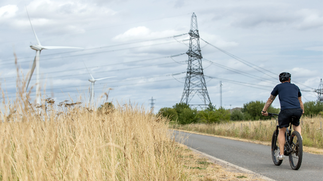 Man wearing t-shirt, shorts and helmet cycling along field towards electricity pylons and wind turbines