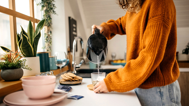 Person wearing orange jumper pouring hot water from a kettle into a cup