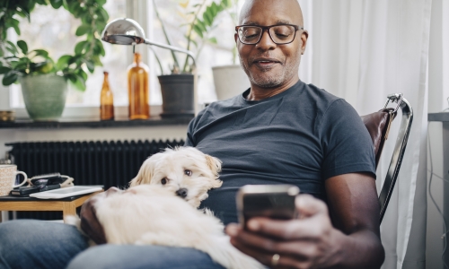 Smiling retired senior male using smart phone while sitting with dog in room at home