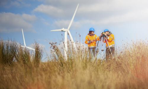 Man and woman standing in grassy field in front of windfarms, wearing hard hats and high-vis jackets with a survey camera - used for the National Grid story 'Now is the time to recruit a Net Zero Energy Workforce to power a greener future'