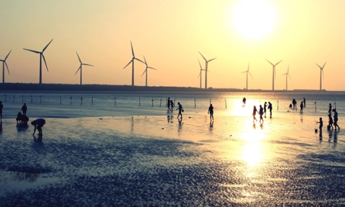 Silhouettes of people on a beach at sunset with wind turbines out at sea in the background
