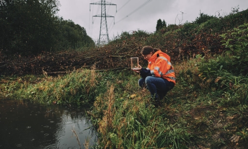 Woman wearing an orange high-vis jacket crouching by a pond and holding a glass jar with a newt