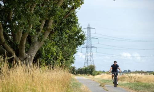 Man wearing t-shirt, shorts and a helmet cycling along a country lane with a pylon in the background