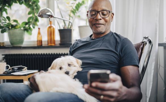 Smiling retired senior male using smart phone while sitting with dog in room at home