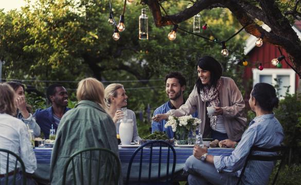 Group of people having dinner in a garden under a tree with a string of lights in it