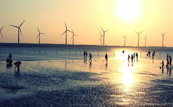 Silhouettes of people on a beach at sunset with wind turbines out at sea in the background