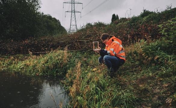 Woman wearing an orange high-vis jacket crouching by a pond and holding a glass jar with a newt