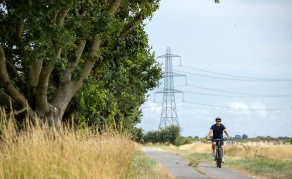 Man wearing t-shirt, shorts and a helmet cycling along a country lane with a pylon in the background