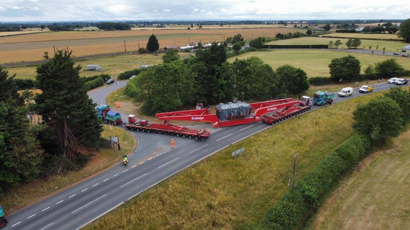 Photo of the delivery. Behind the large delivery vehicle, there is a convoy of other vehicles.