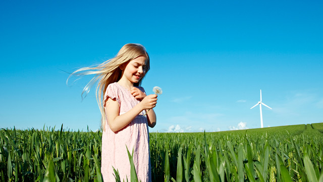 Fille tenant une horloge en forme de pissenlit dans un champ avec une éolienne en arrière-plan.