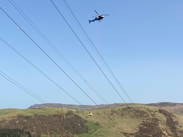 Helicopter hovering over electricity overhead lines with basked carrying engineers suspended below