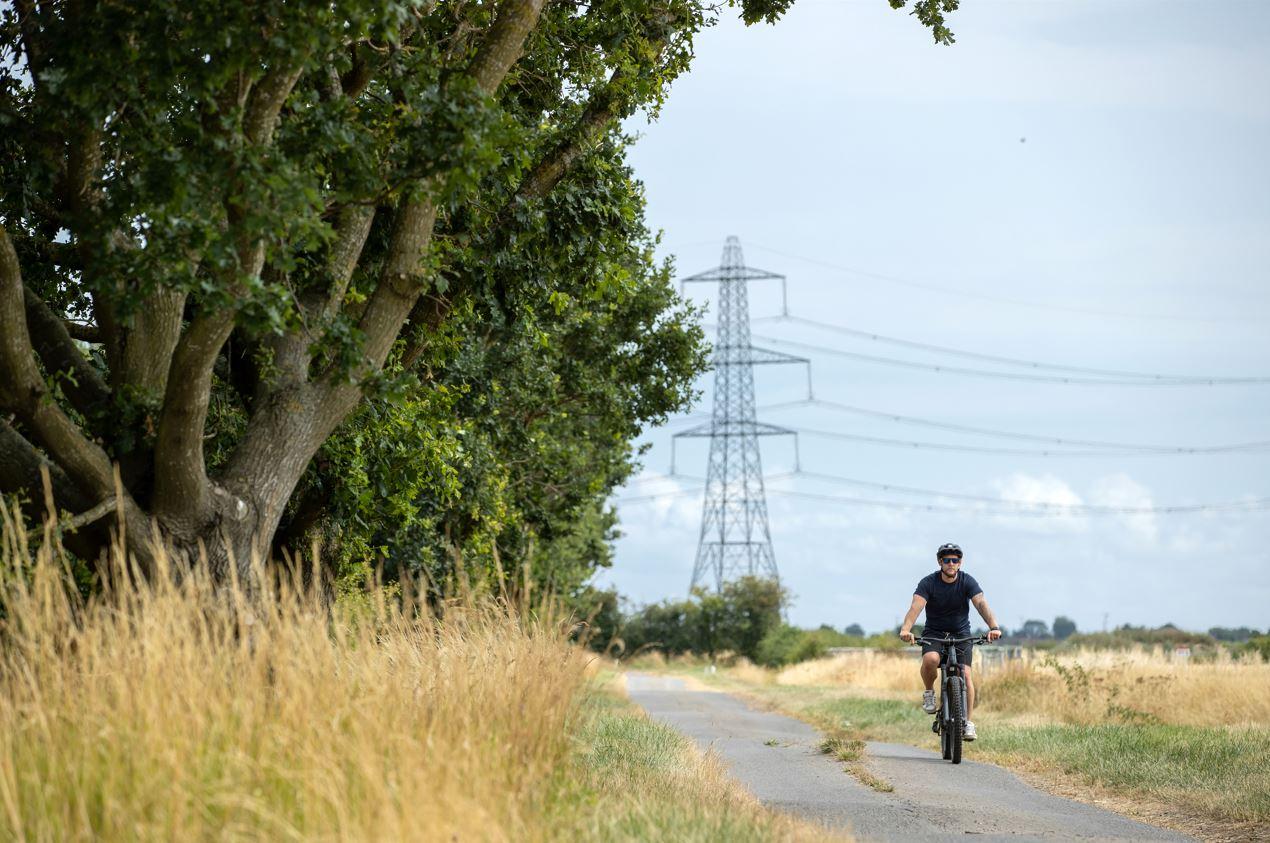 Bicker Fen Cyclist.jpg Man wearing t-shirt, shorts and a helmet cycling along a country lane with a pylon in the background