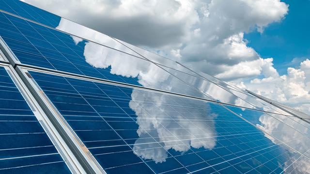 Solar panels beneath a blue, cloudy sky with the clouds reflecting in the panels