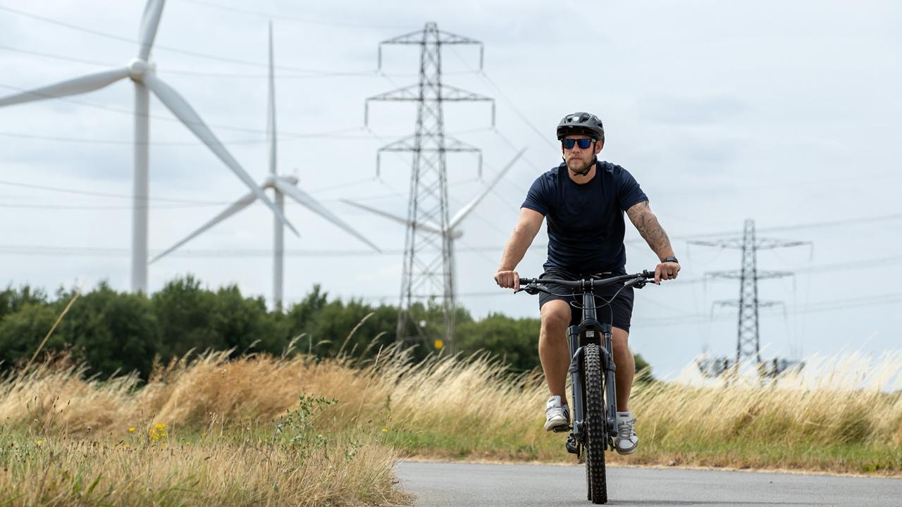 CleanEnergyTransition_BickerFen_1280x720.jpg Man wearing t-shirt, shorts and a helmet cycling along a country lane in front of wind turbines and pylons