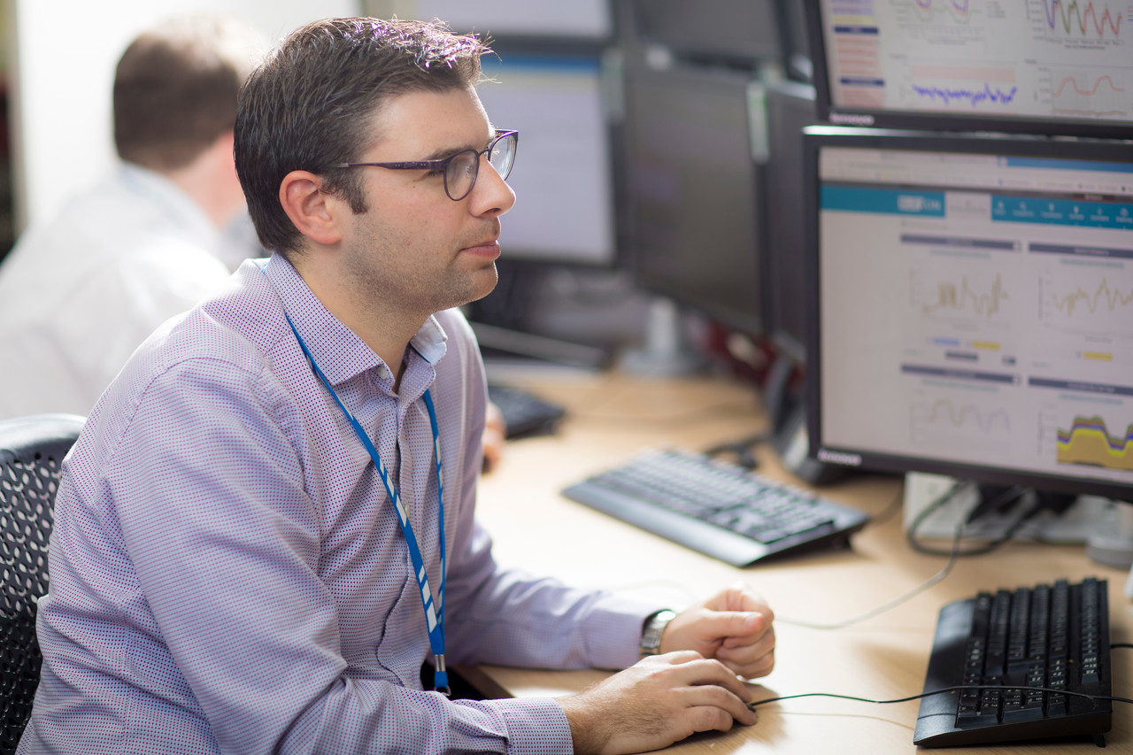 Man wearing glasses and purple shirt and a lanyard looking at screens in a control centre