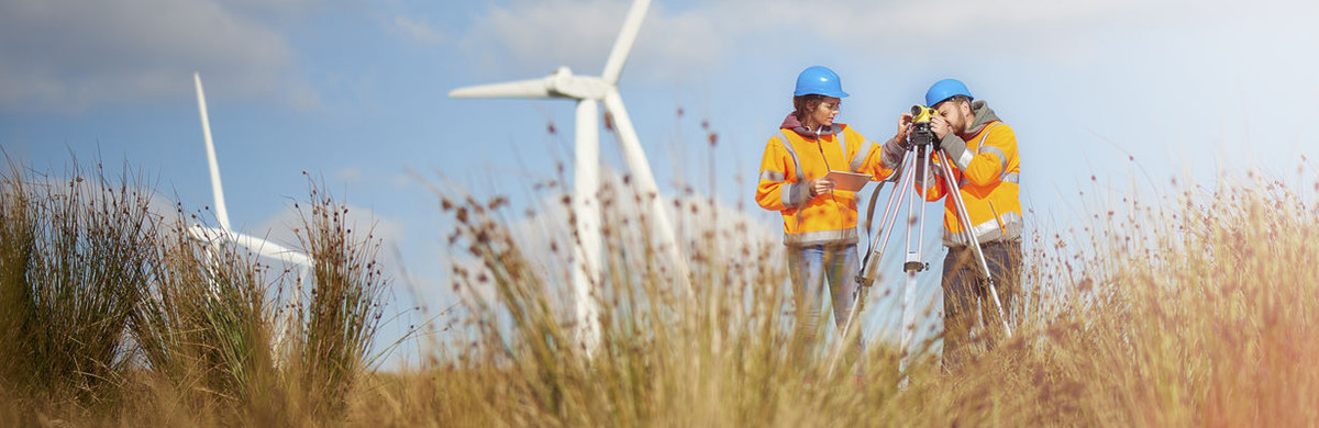 Man and woman standing in grassy field in front of windfarms, wearing hard hats and high-vis jackets with a survey camera - used for the National Grid story 'Now is the time to recruit a Net Zero Energy Workforce to power a greener future'