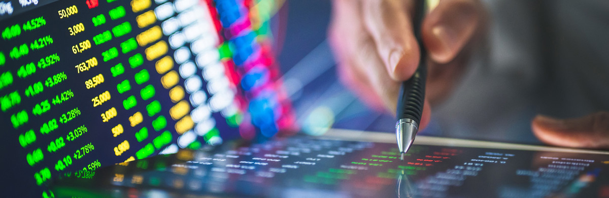 Hand holding a pen working on a smart-pad with stock market trading screens to the side