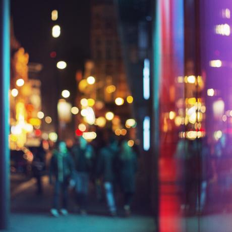 An out of focus city view with well-lit buildings and people walking along the pavement 