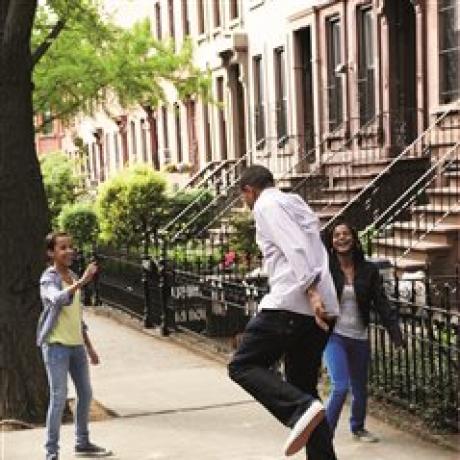 A family of three dancing on a sidewalk with buildings and a tree in the background 