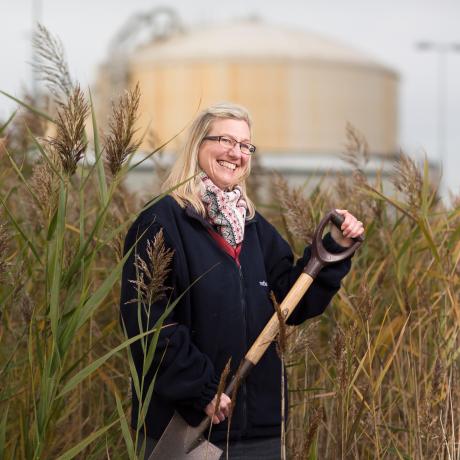 A National Grid representative standing in a field of high reeds holding a shovel with an industrial building in the background 