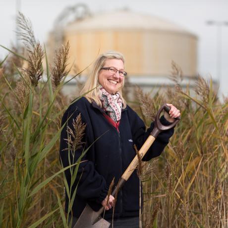 A National Grid representative standing in a field of high reeds holding a shovel with an industrial building in the background