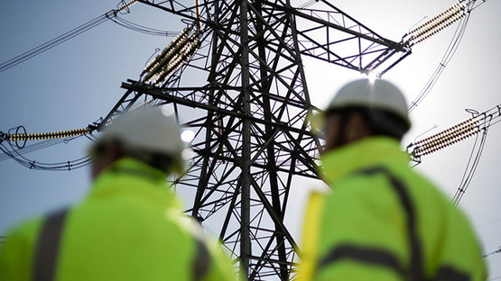 Two people wearing high-vis jackets and hard hats standing in front of electricity pylon