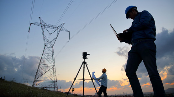 Two engineers wearing hard hats surveying land next to electricity pylon at dusk